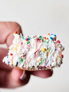 a hand holding a piece of cake with white frosting and sprinkles