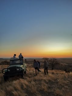 three people standing next to a vehicle in the middle of a dry grass field at sunset