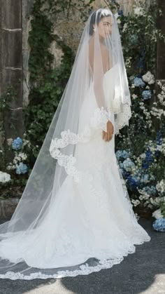 a woman in a wedding dress and veil is standing by some flowers with her back to the camera