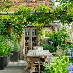 an outdoor dining table surrounded by greenery and potted plants in front of a brick building