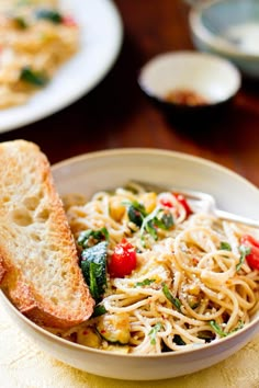 a bowl filled with pasta and veggies on top of a wooden table next to two slices of bread