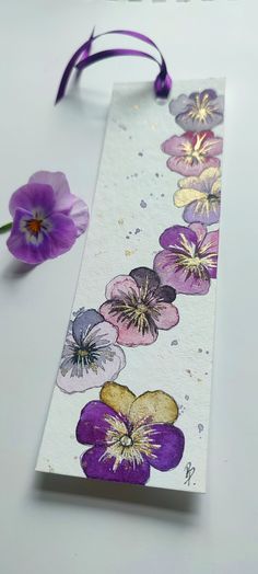 a bookmark with purple and yellow flowers painted on it, next to a flower