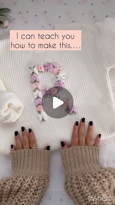 a woman's hands with black nail polish holding up a white knitted sweater that says i can teach you how to make this