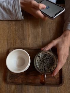 a person holding a cell phone over a cup on a wooden tray next to a coffee mug