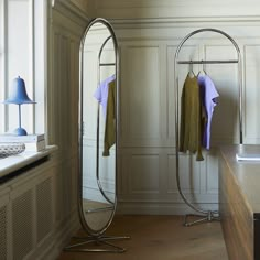 an oval shaped mirror in the corner of a room with clothes on hangers and a lamp next to it