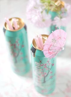 three green cans with pink umbrellas and flowers in them on a tableclothed surface