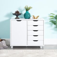 a white cabinet with five drawers in front of a blue wall and potted plants