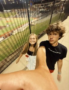 two young people standing next to each other in front of a fence and baseball field