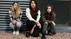 three young women sitting on the ground in front of a metal fence, one with her hand on her hip