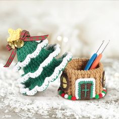 two crocheted christmas decorations are sitting on the snow covered ground, one has a small house with a green roof and another has a red ribbon