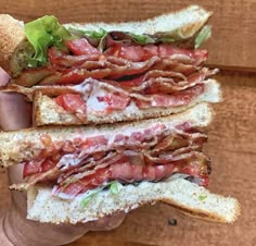 a hand holding a sandwich with bacon, lettuce and tomato on it in front of a wooden wall