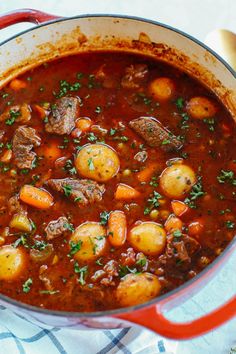 a red pot filled with stew and potatoes on top of a blue towel next to bread