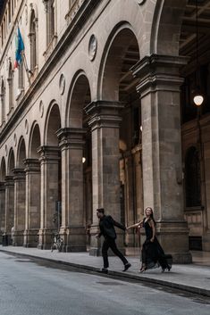 two people walking down the street in front of an old building with arches and pillars