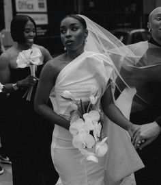 a woman in a wedding dress standing next to two other women on the street with flowers