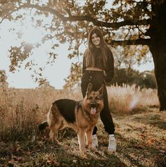 a woman standing next to a dog on top of a grass covered field with trees in the background