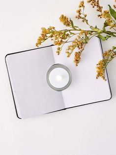 an open book with a lit candle and flowers on the cover next to it, sitting on a white surface