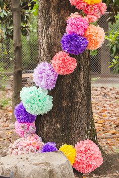 colorful paper flowers hanging from a tree in front of a rock and some leaves on the ground