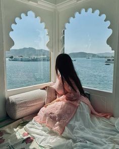 a woman sitting on top of a bed next to a window looking out at the water