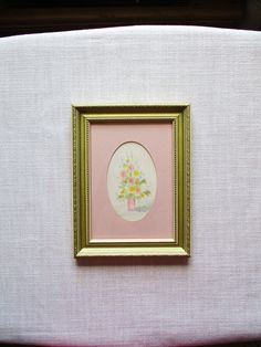 a pink and gold frame with a flower arrangement in it on a white surface next to a pillow