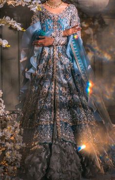 a woman in a blue and silver dress with flowers around her neck, standing next to a tree