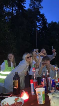 a group of people standing around a table with drinks and food on it at night