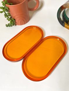 two orange plates sitting on top of a white table next to a cup and plant