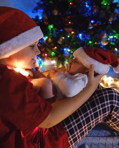 a woman holding a baby wearing a santa hat while sitting in front of a christmas tree