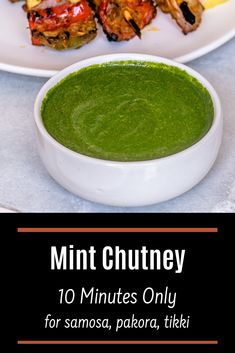 a white plate topped with food next to a bowl of green soup and chicken skewers