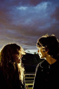 two people standing next to each other in front of a sky filled with dark clouds