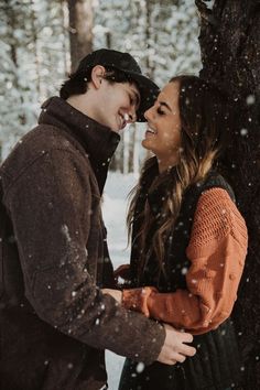 a man and woman standing next to each other near a tree in the snow with their faces close together