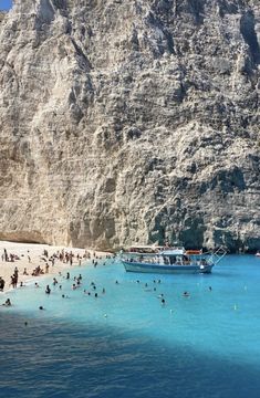 many people are swimming in the blue water near a rocky cliff and boat on the beach