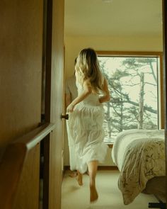 a woman in a white dress is walking into a room with a bed and window