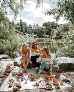 three women sitting on a blanket near a river eating food and drinking watermelon