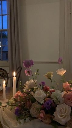 a table topped with a vase filled with flowers next to two lit candles and a window