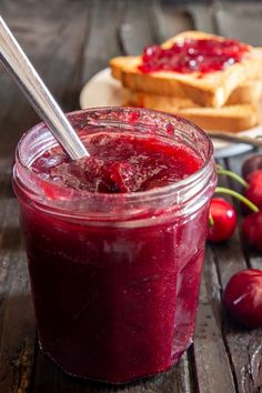 a glass jar filled with liquid next to some cherries on a wooden table and bread in the background