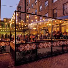 people are sitting at tables in an outdoor area with string lights strung over the buildings