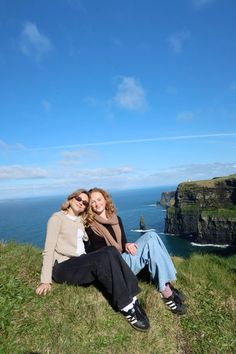 two women sitting on top of a grass covered hill next to the ocean with cliffs in the background