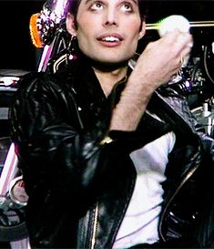 a man in black leather jacket holding a white object up to his face while standing next to a motorcycle