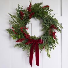 a wreath with red berries and green leaves hanging on a white door, next to a red ribbon