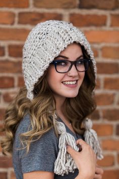 a young woman wearing glasses and a knitted hat smiles while standing in front of a brick wall