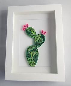 a green and pink paper art piece in a white frame on a gray surface with two small pink flowers