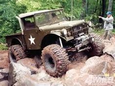 a man standing next to a green jeep on top of a pile of rocks in the woods