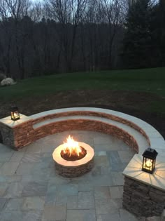 an outdoor fire pit surrounded by stone walls and lanterns at dusk with trees in the background