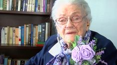 an old woman with glasses holding flowers in front of a book shelf filled with books