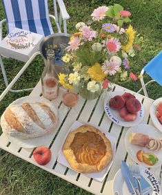 a table with bread, fruit and other food on it in the grass next to flowers