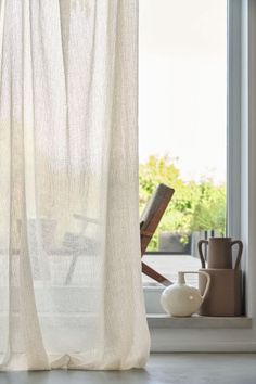 an open window with white curtains and vases on the floor in front of it