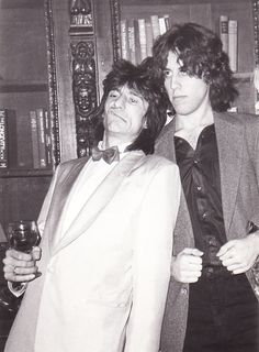 two young men standing next to each other in front of bookshelves holding wine glasses