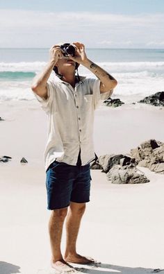 a man standing on top of a sandy beach next to the ocean holding a camera