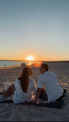 a man and woman sitting on the beach at sunset