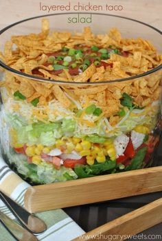 a salad in a glass bowl with wooden utensils on the side and text overlay that reads layered chicken taco salad